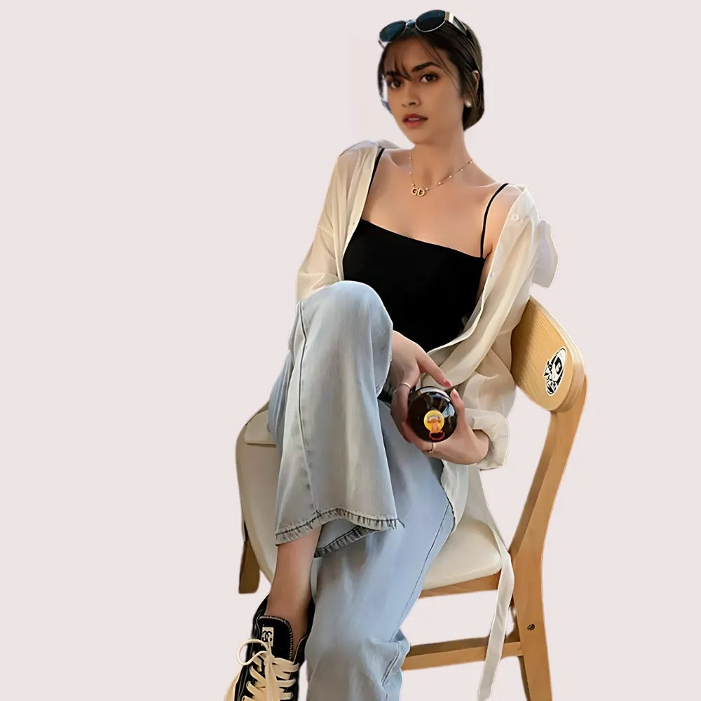 Woman sitting on a chair holding a bottle, wearing a black top and light blue jeans, with a neutral background.