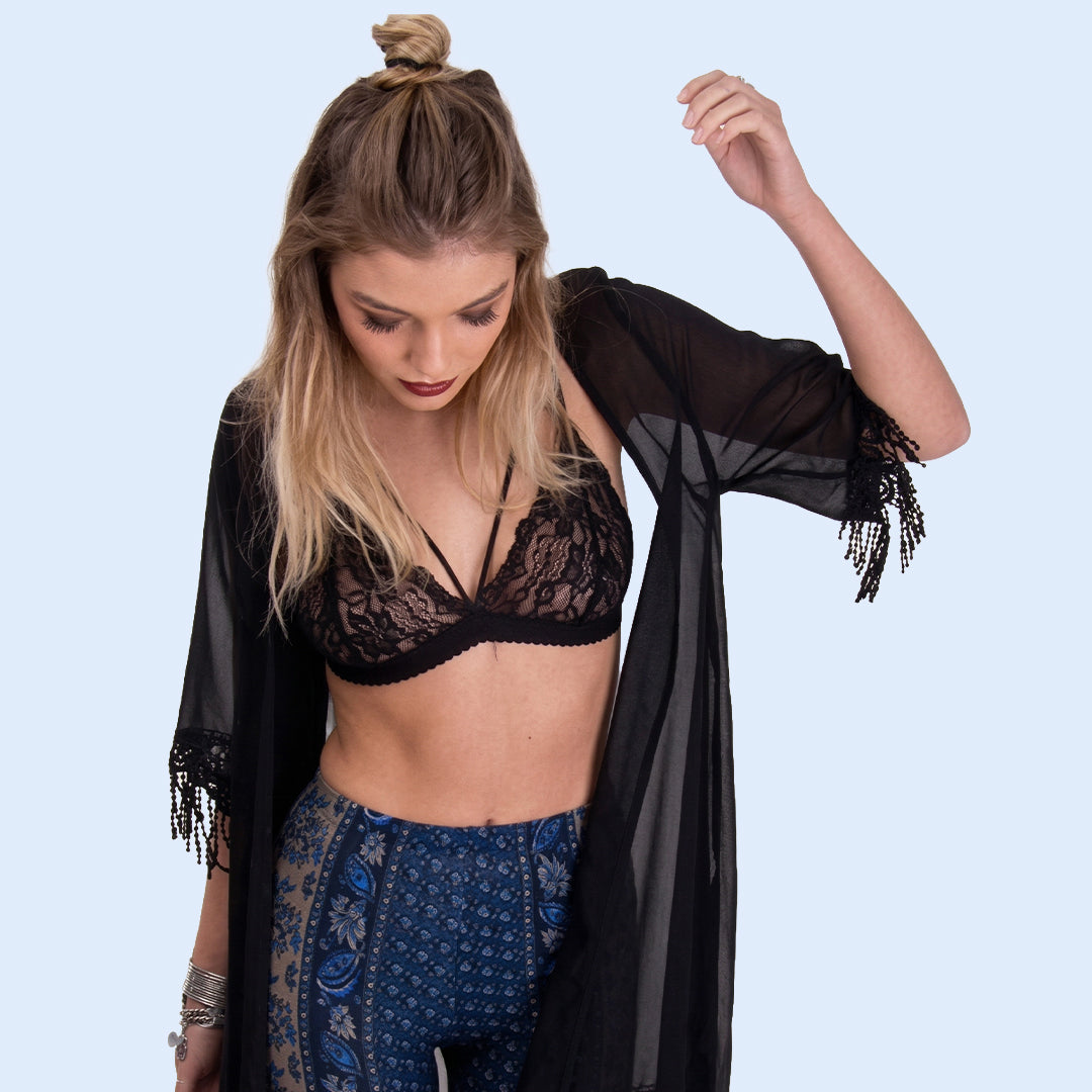 Woman wearing a black lace bralette and blue patterned pants with a black sheer shawl on a light blue background