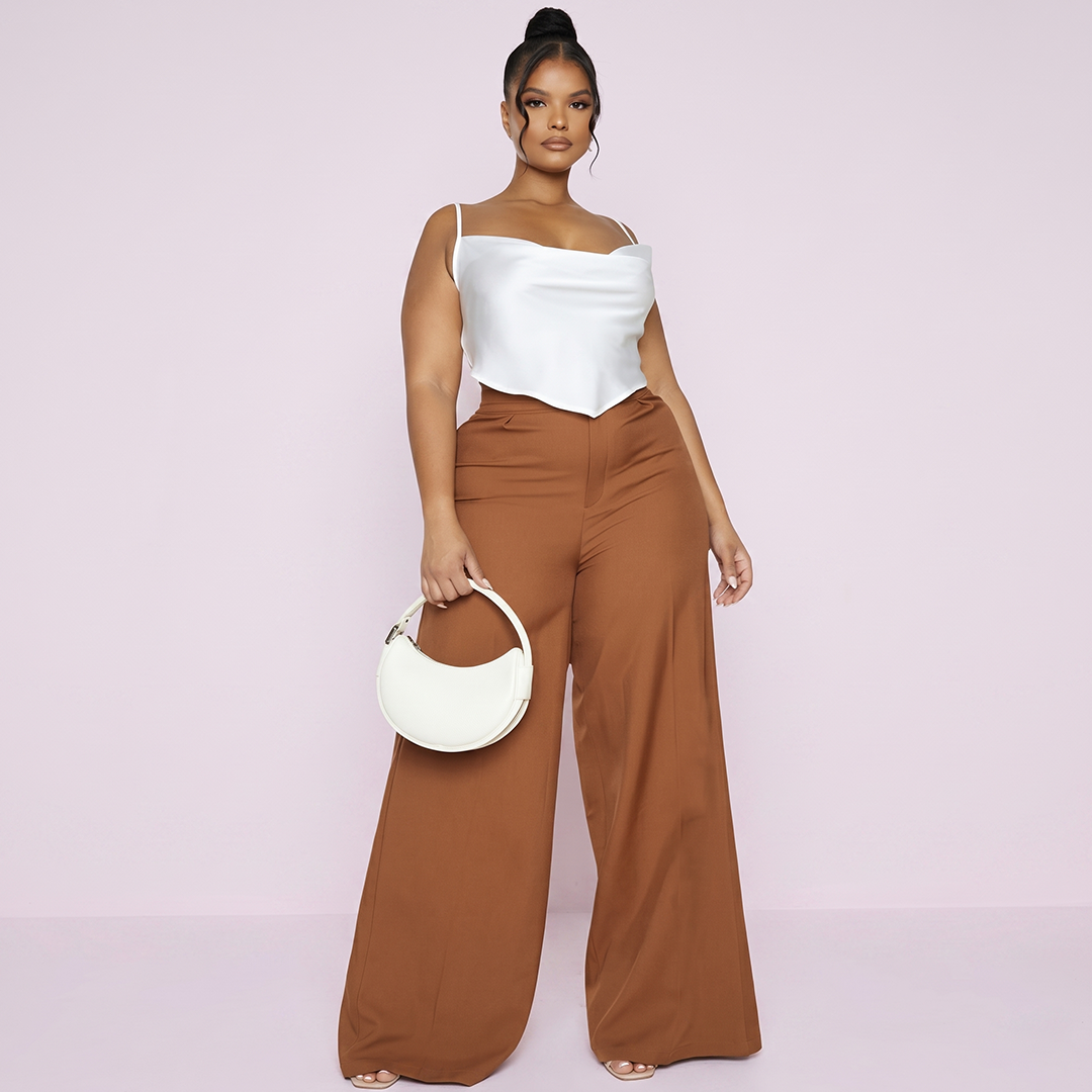 Woman wearing a white top and brown pants holding a white handbag on a light pink background